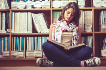 Female student against bookshelf reading a book on the library f