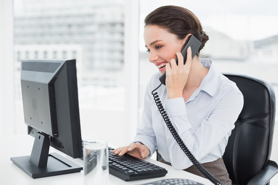 Smiling Businesswoman Using Landline Phone And Computer In Offic