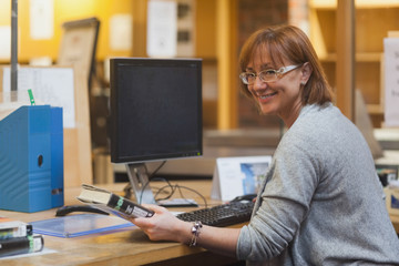 Smiling female librarian holding a book standing behind the desk - obrazy, fototapety, plakaty