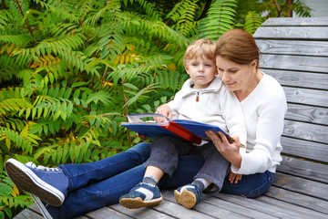 Little boy and his mother sitting on bench in park and reading b