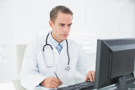 Doctor writing a note while using computer at medical office
