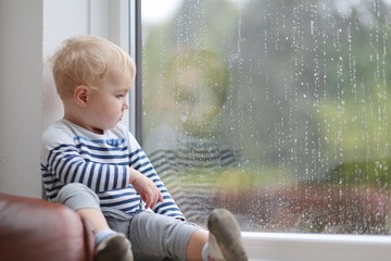 Cute baby girl looking outside through the window during rain