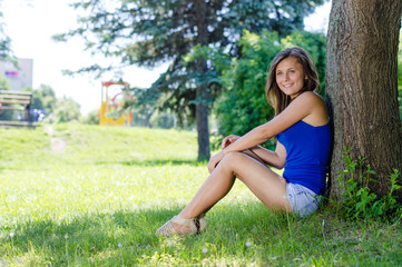 Naklejka na ściany i meble Happy school girl sitting at tree in park on summer day
