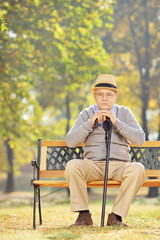 Thoughtful senior man with a cane sitting on a bench in a park