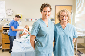 Confident Nurses Standing Against Couple With Baby In Background