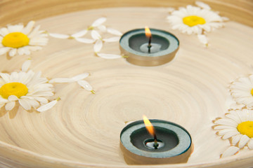 Two burning candles, heads of camomiles in dish with water