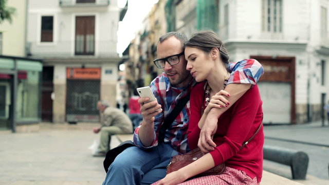 Young couple sitting on bench in city and watching on smartphone