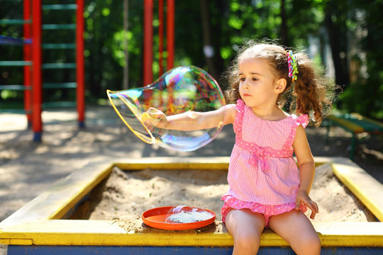 Little girl with tails sitting in a sandbox and blows bubbles