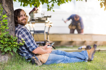 Carpenter Holding Mobilephone And Cup While Leaning On Tree Trun