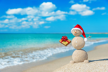 Smiley sandy snowman at beach in christmas hat with golden gift.