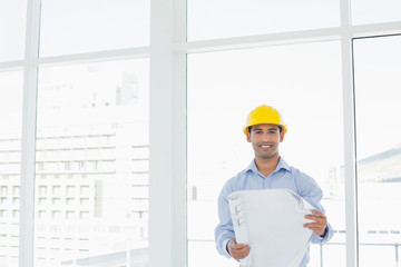 Smiling architect in yellow hard hat with blueprint in office