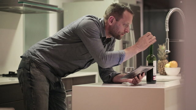 Young man with tablet computer drinking wine in kitchen