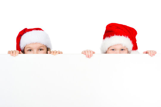 Two little kids looking over white poster in red Santa hats.
