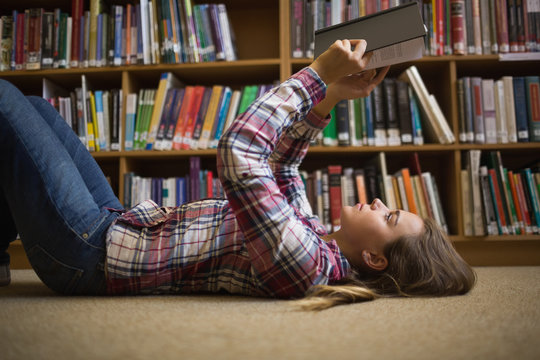 Pretty student lying on library floor reading book