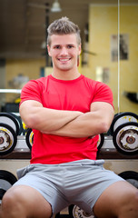 Fototapeta na wymiar Handsome smiling young man in gym sitting on dumbbells rack
