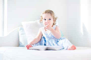 Cute toddler girl in a blue dress with curly hair reading a book