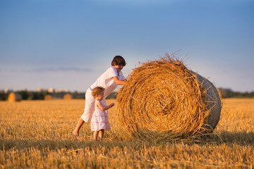 Naklejka na ściany i meble Brother and baby sister pushing hay bales in a field at sunset