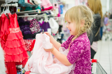 Adorable girl on shoppping cart select pink dress in supermarket