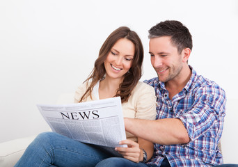 Happy Young Couple Sitting On Couch Reading Newspaper