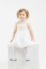 Little girl in white dress sits on big cube on white background.