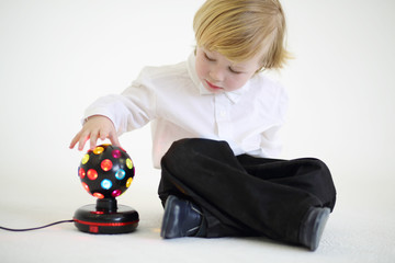 Little blond boy sits on floor with ball-shaped lamp on white