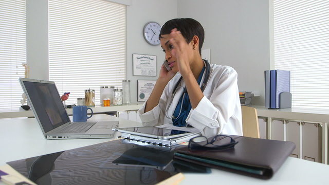 Black doctor at desk talking on smartphone and looking at laptop