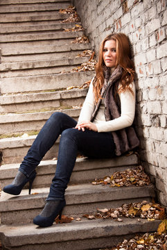 slim woman in jeans sitting on stairs covered with yellow leaves