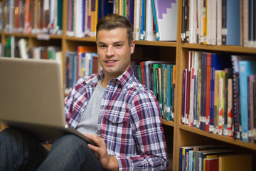 Smiling young student sitting on library floor using laptop