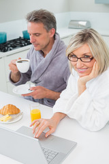 Couple in bathrobes with coffee and juice using laptop in kitche