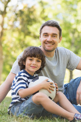 Naklejka premium Father and son with football sitting on grass inpark
