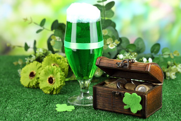 Glass of green beer and pitcher with coins on grass close-up