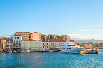 Obraz premium A large luxury yacht rests at the harbor of a historic Mediterranean town, featuring traditional stone architecture and warm sunlight reflecting off the calm blue sea under a clear sky.