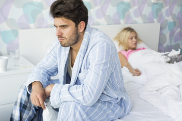 Obraz premium Sick man sitting on bed with woman sleeping in background at home
