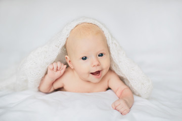Happy baby lying on white bed under soft blanket, smiling and looking at camera