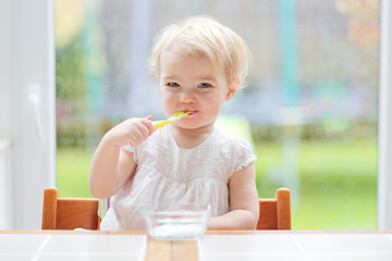 Happy toddler girl eating yogurt sitting next to big window