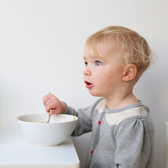 Sweet blonde toddler girl eating porridge with spoon 
