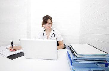 Young female doctor with laptop writing notes on desk in clinic