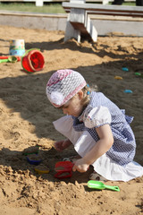 Little girl in dress plays in sandbox with shovel on summer day.