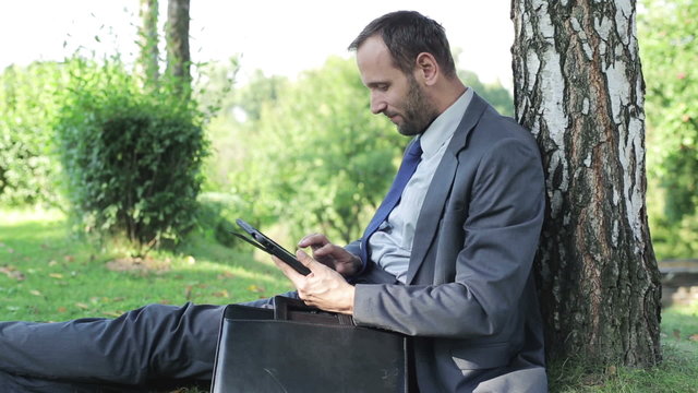 businessman sitting under the tree shadow and working on his tab