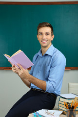 Young teacher sitting with book on desk in school classroom