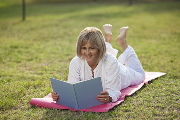 Naklejka na ściany i meble senior woman reading in park laying on yoga mat
