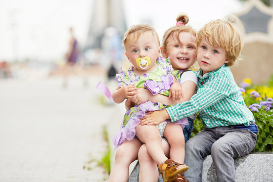 Three Children Sit Embracing On Granite Curb Of Flowerbed