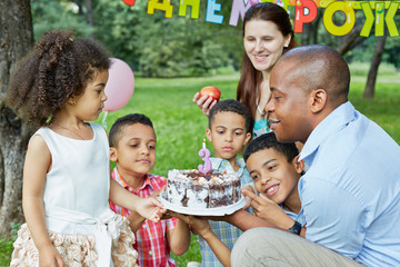 Family of six looks at birthday cake with burning candle on it