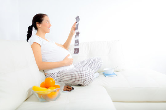 Beautiful pregnant woman with baby photos on sofa at home