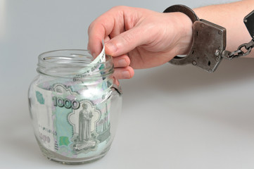 Рука в наручниках берет деньги из стеклянной банки