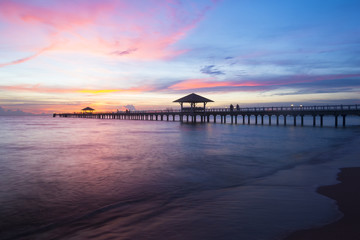 Fototapeta premium Wooden pier at Casalunar Bangsaen Village, Chonburi, Thailand, extending across calm sea during colorful sunset, featuring pavilion, dramatic twilight sky, peaceful coastline, tropical travel.