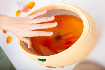 Female hand and orange paraffin wax bowl. Woman in beauty salon - obrazy, fototapety, plakaty