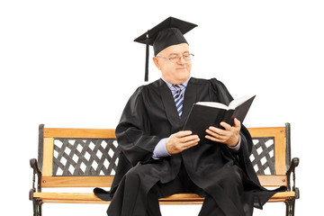 Mature man in graduation gown seated on bench reading book