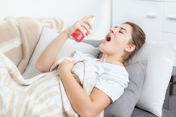 Obraz na płótnie Canvas Young woman using throat spray while lying on couch