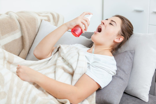 Young sick woman lying in living room and using throat spray